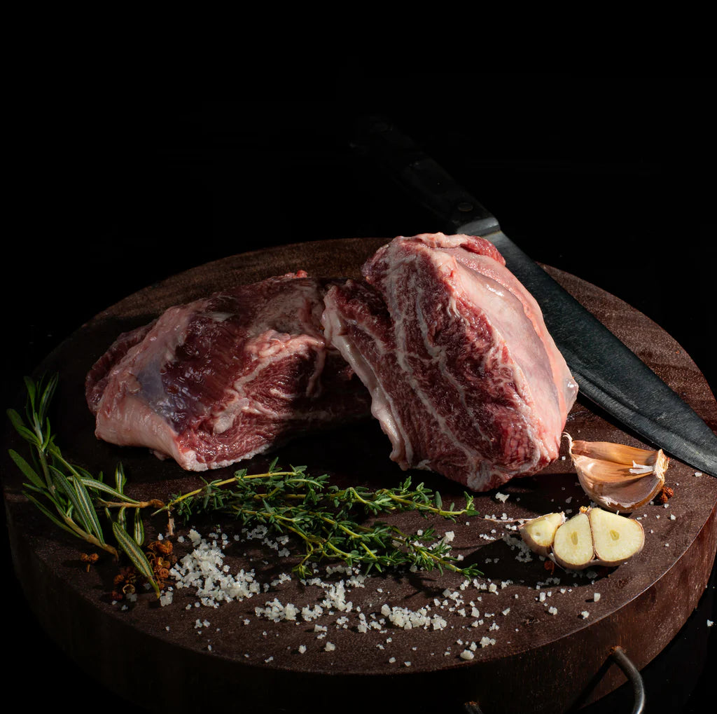 Black Onyx Beef Cheeks MS 3+ on a cutting board with rosemary, thyme, garlic, salt, and a knife.