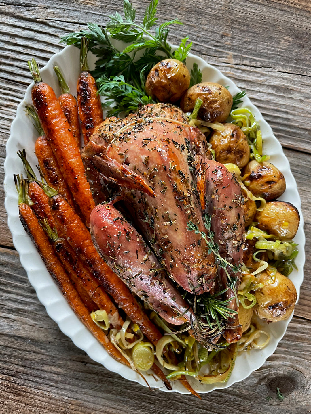 Whole roasted pheasant garnished with herbs, served with roasted carrots and potatoes on a white platter.