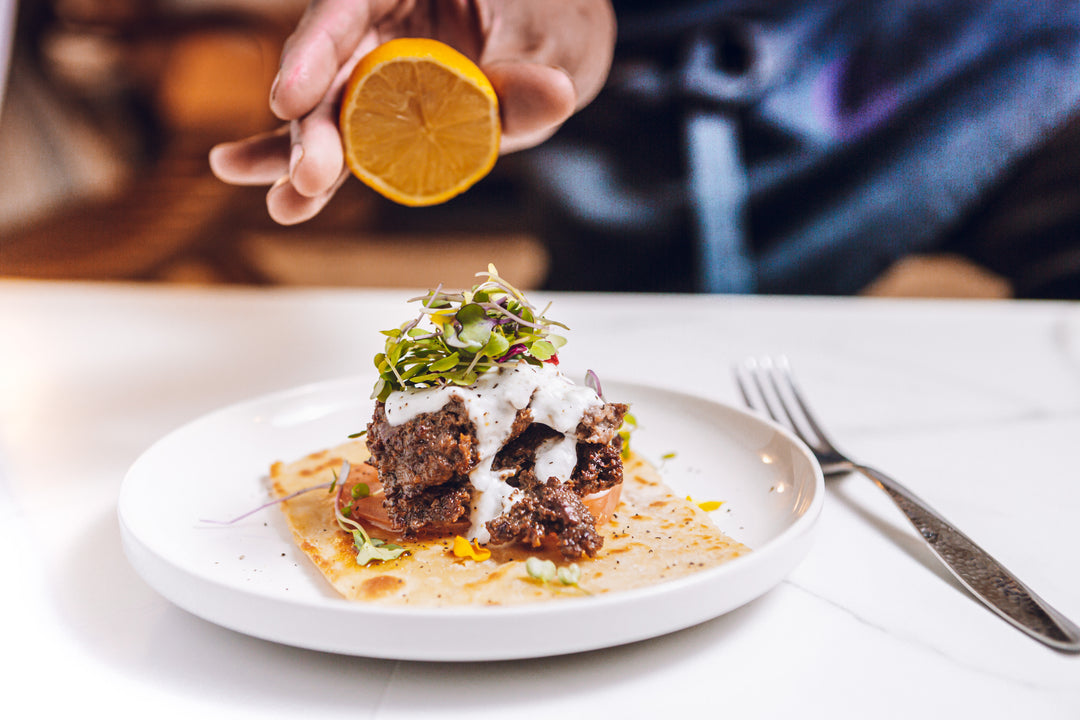 A delicious lamb burger topped with microgreens and sauce, served on flatbread with lemon, on a white plate.
