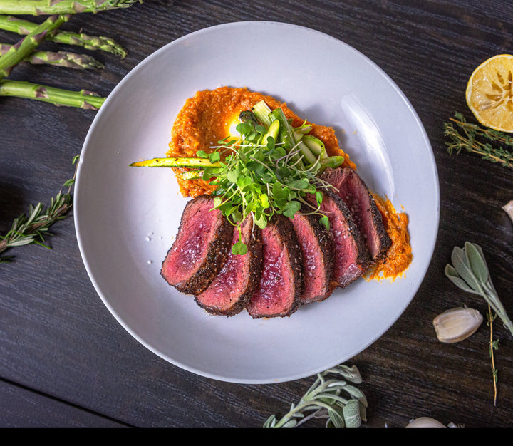 Sliced bison NY strip steak served with creamy red pepper sauce and topped with microgreens.