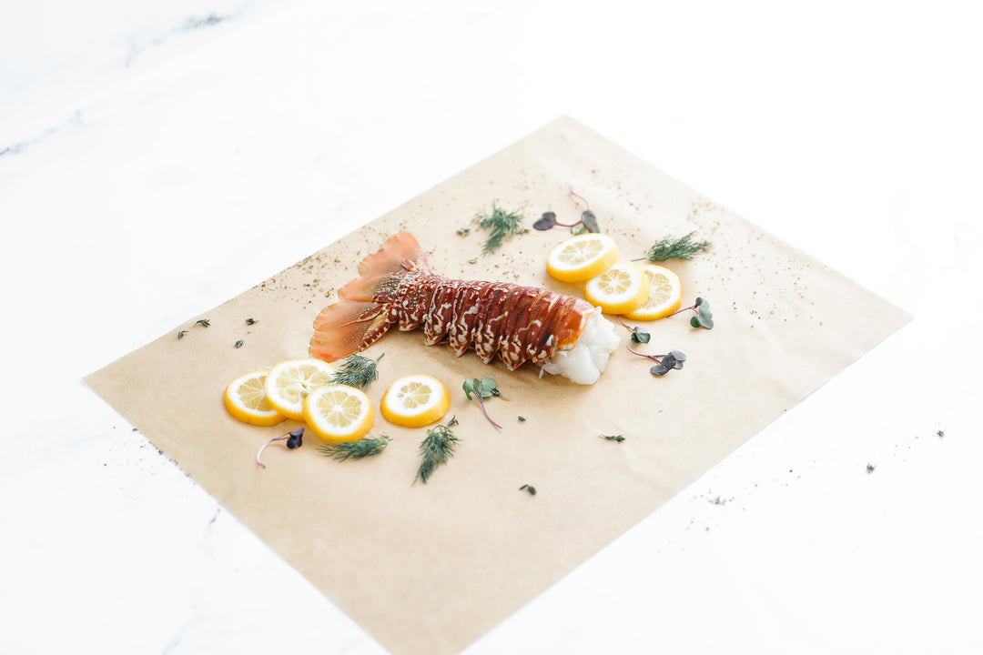 Raw lobster tail on parchment with lemon and garnishes, ready for cooking.