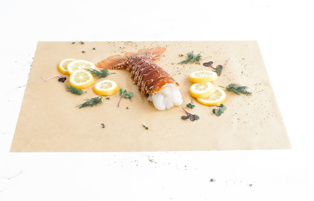 South African lobster tail on parchment surrounded by lemon slices and herbs.