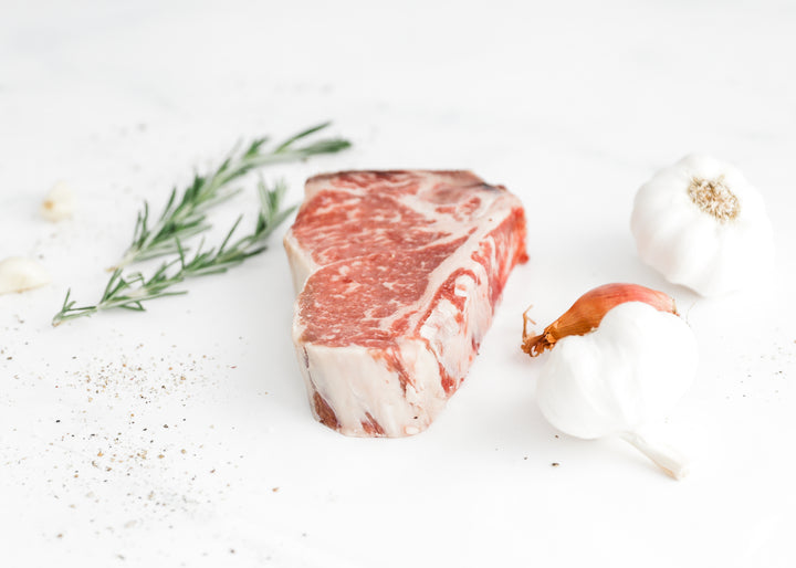 Close-up of a raw, marbled USDA Prime Bone-In New York Strip steak with herbs.