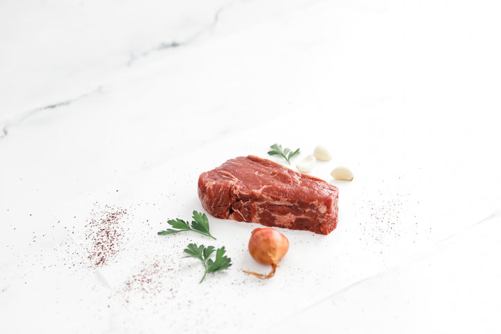 A fresh bison NY strip steak, garnished with herbs and garlic on a white surface.