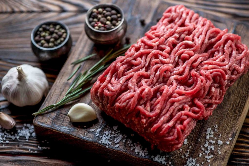 Fresh ground elk meat on a wooden board, garnished with garlic, herbs, and pepper.