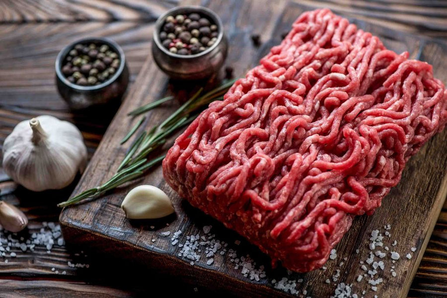 Fresh ground elk meat on a wooden board, garnished with garlic, herbs, and pepper.