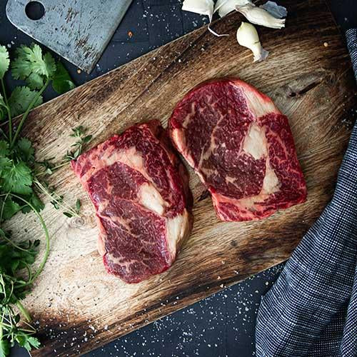 Two Grass-Fed Angus PureNZ Boneless Ribeye steaks on a wooden board with herbs and garlic, ready to cook.